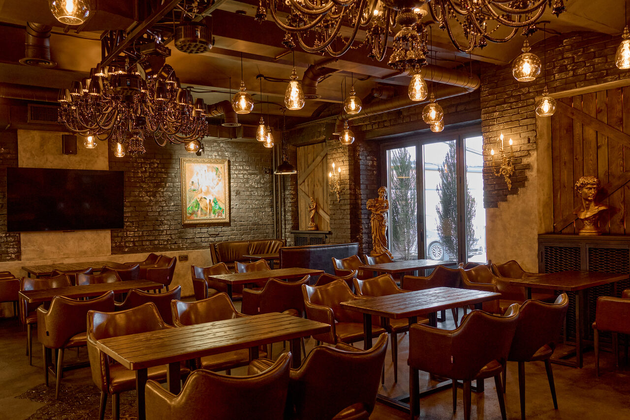 Арендовать Tangiers Lounge(Танжер Лаундж) . Пространство с кирпичными стенами, несколько деревянных столов и кожаных стульев, крупная люстра и подвесные лампы, окно с растениями снаружи, картина на стене, декоративные бюсты, металлические элементы интерьера, коричнево-бежевые тона.