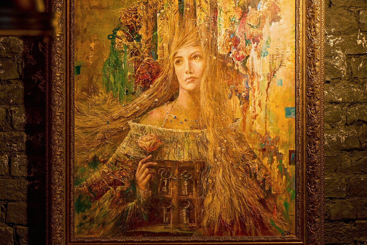 Арендовать Tangiers Lounge(Танжер Лаундж) . Картина в золотистой раме на кирпичной стене. Изображение женщины с длинными волосами, держащей розу и архитектурные элементы. Преобладают теплые оттенки зеленого и оранжевого. Освещение мягкое, акцент на картине.