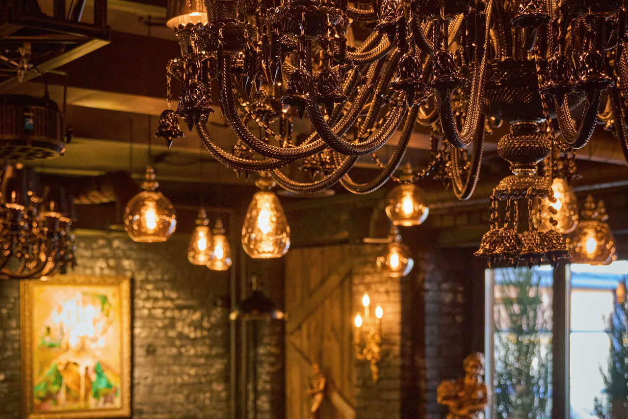 Арендовать Tangiers Lounge(Танжер Лаундж) . Лофт-пространство с кирпичными стенами темного цвета, украшенное массивными черными люстрами и подвесными лампами теплого света. Видно деревянную дверь, настенное искусство в раме и окна с естественным освещением.