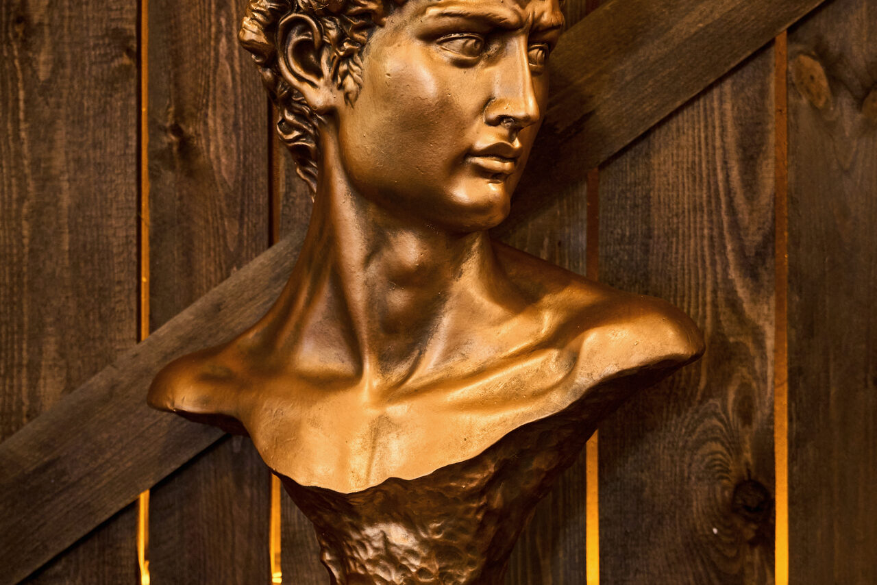 Арендовать Tangiers Lounge(Танжер Лаундж) . Бюст мужчины в античном стиле из бронзового материала на фоне деревянной стены с диагональными панелями. Освещение тёплое, подчёркивающее текстуру дерева и детали бюста. Дерево имеет тёмный оттенок и вертикальные линии текстуры.