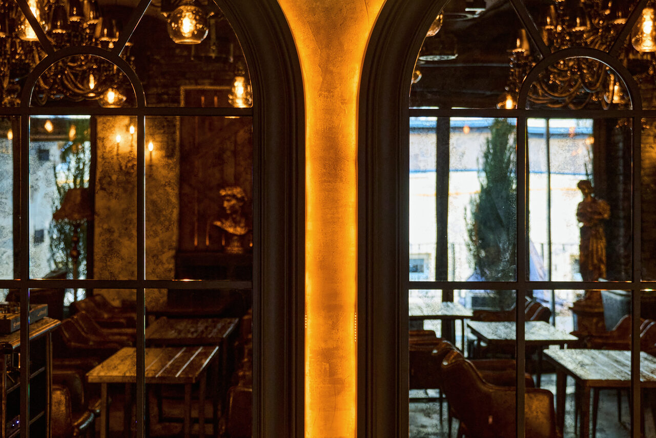 Арендовать Tangiers Lounge(Танжер Лаундж) . Интерьер ресторана с деревянными столами и стульями, стены из кирпича, теплое освещение от множества подвесных светильников, стеклянные окна с арочными рамами, декоративные статуи, темные тона, уютная атмосфера.