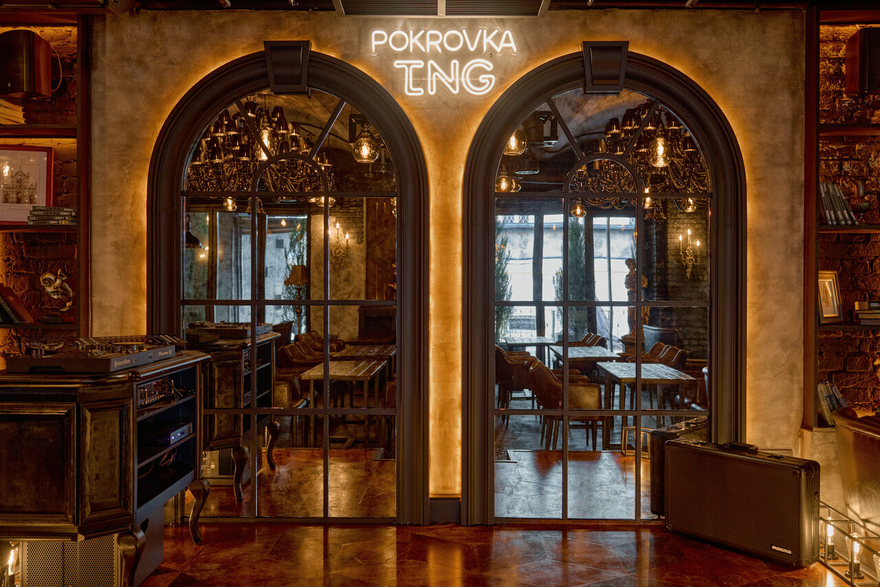 Арендовать Tangiers Lounge(Танжер Лаундж) . Арочные стеклянные двери с металлическими рамами, за ними видны столы и стулья. На стене над дверями светится неоновая надпись "POKROVKA ING". Потолочные светильники в стиле ретро. Теплая цветовая гамма с элементами древесных текстур.