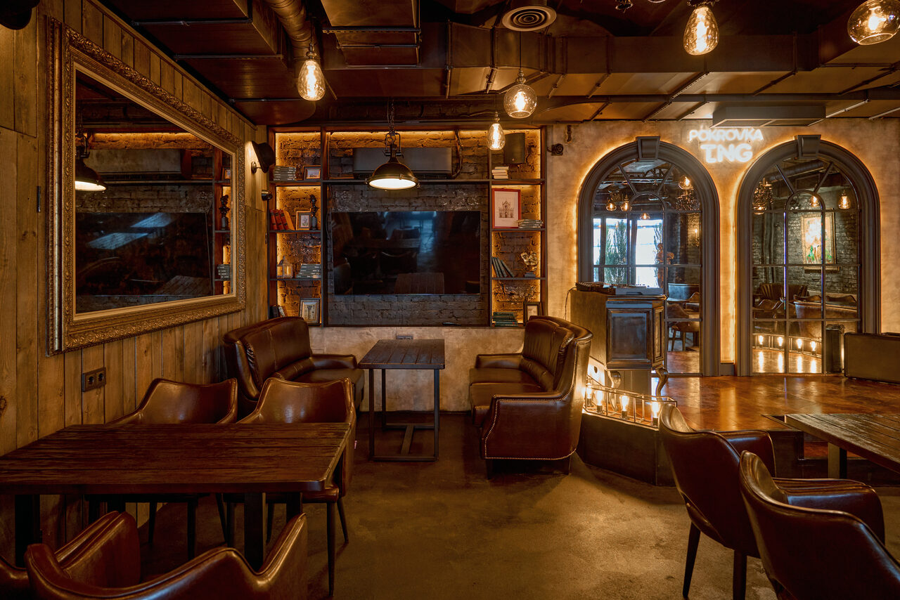 Арендовать Tangiers Lounge(Танжер Лаундж) . Бар с деревянными стенами и потолком, кожаными креслами и столами. На стене крупное зеркало, открытки и книги на полках. На потолке подвесные лампы с теплым светом. Над аркой неоновая надпись "PENROVA TING". Пол бетонный.