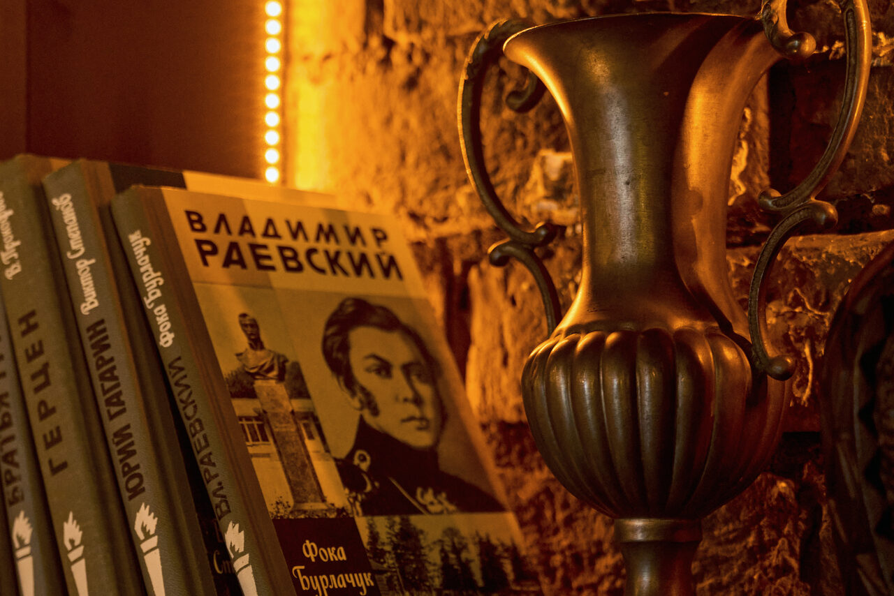 Арендовать Tangiers Lounge(Танжер Лаундж) . Книжная полка с книгами, включающими том с именем Владимир Раевский на корешке и обложке. Рядом стоит бронзовая ваза с узорами. Кирпичная стена на заднем плане, освещенная теплым светом.