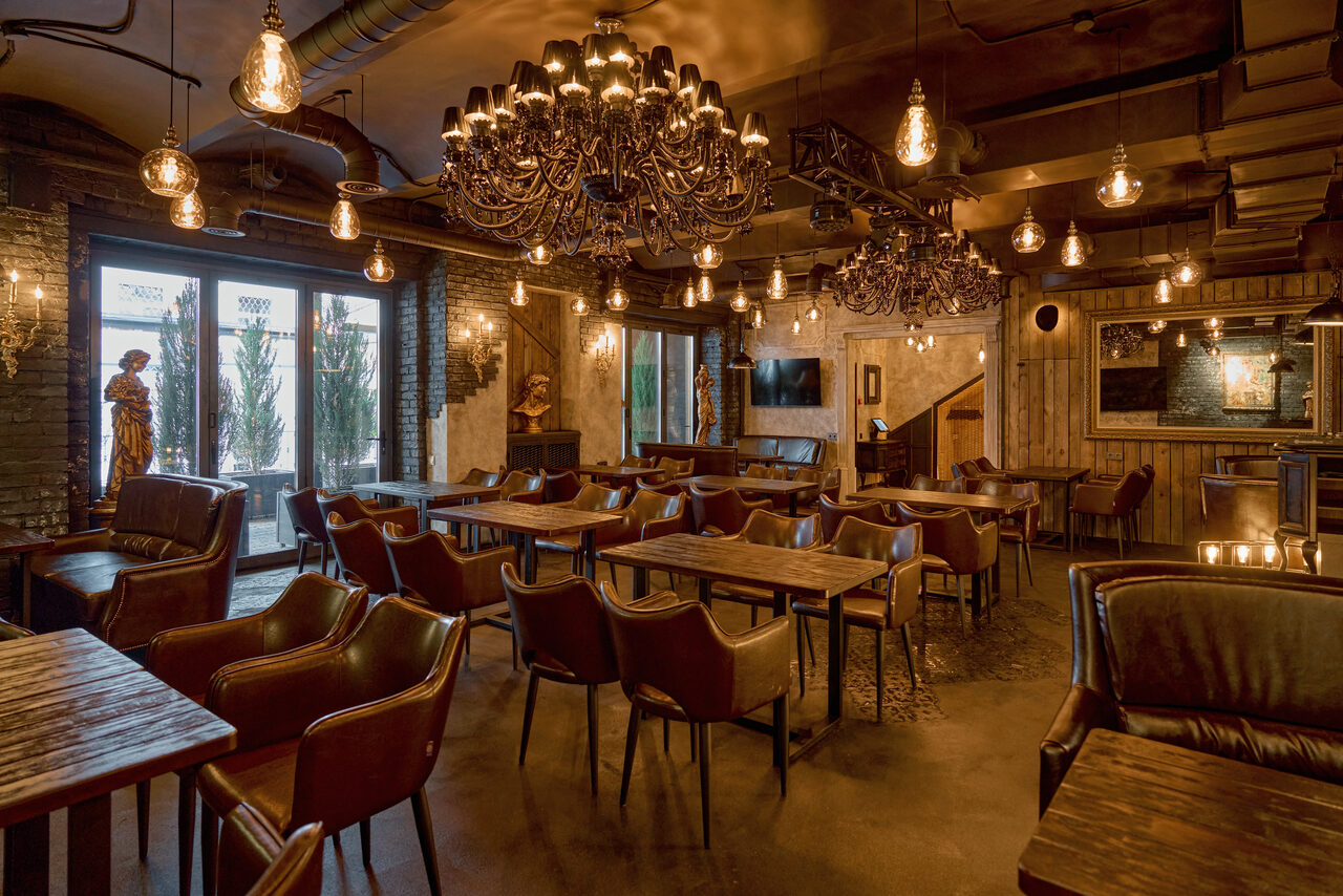 Арендовать Tangiers Lounge(Танжер Лаундж) . Лофт-пространство с деревянными столами и кожаными креслами. Подвесные лампы и люстры. Кирпичные стены, высокие окна с растениями. Телевизор на стене, металлические элементы декора. Теплое освещение, темные и светлые тона.