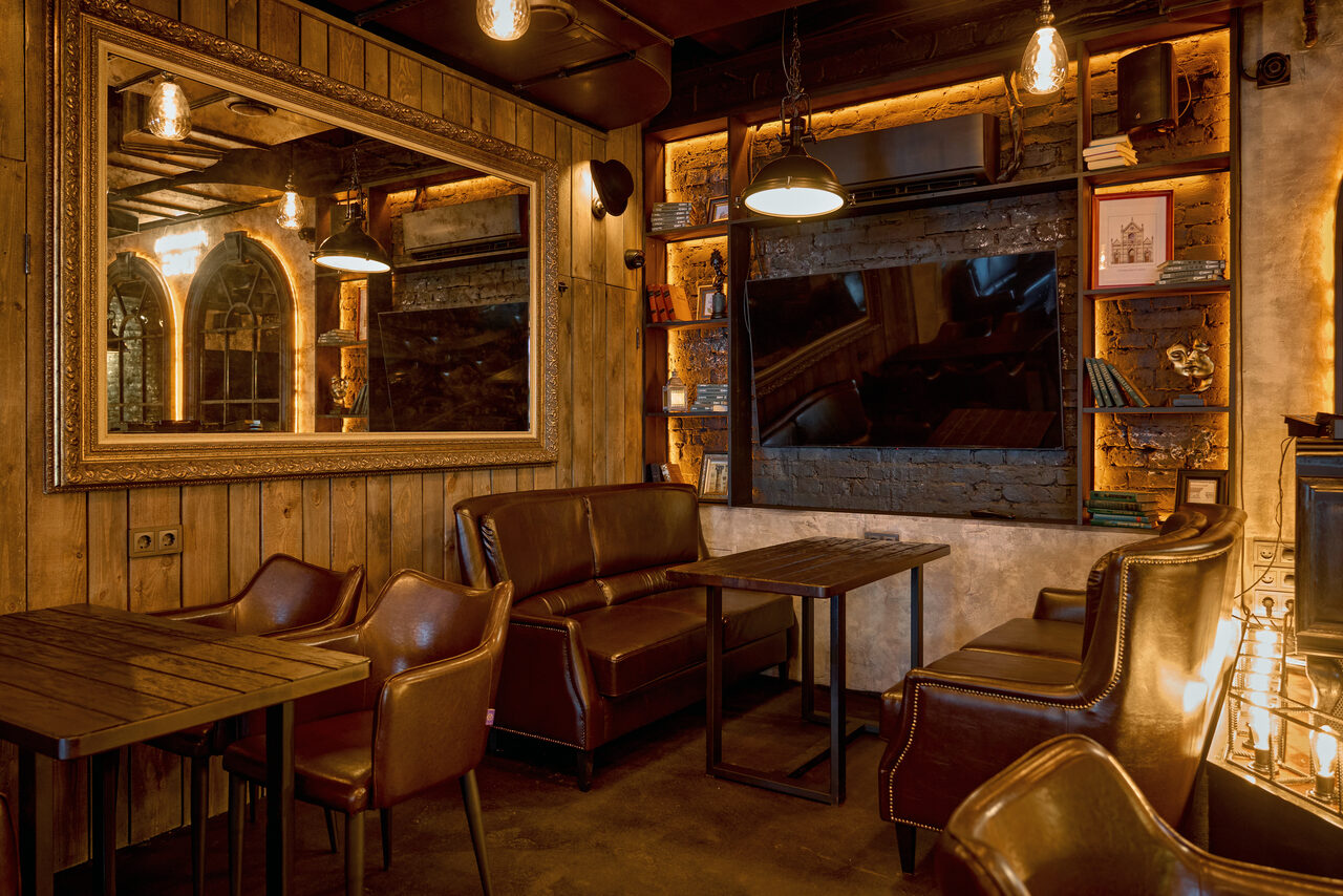 Арендовать Tangiers Lounge(Танжер Лаундж) . Лофт-пространство с деревянными стенами и потолком, коричневыми кожаными диванами и креслами, темными деревянными столами. На стене большое зеркало и полки с книгами. Мягкое освещение, подвесные лампы, кирпичная отделка, теплые коричневые и бежевые тона.