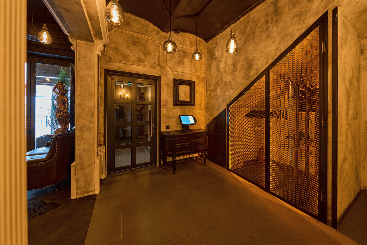 Арендовать Tangiers Lounge(Танжер Лаундж) . Интерьерное пространство с коричневыми стенами в стиле лофт, стеклянная дверь, низкое освещение с подвесными лампами, деревянная мебель, металлическая перегородка, пол темный, присутствует оборудование на столе, зеркальная колонна.