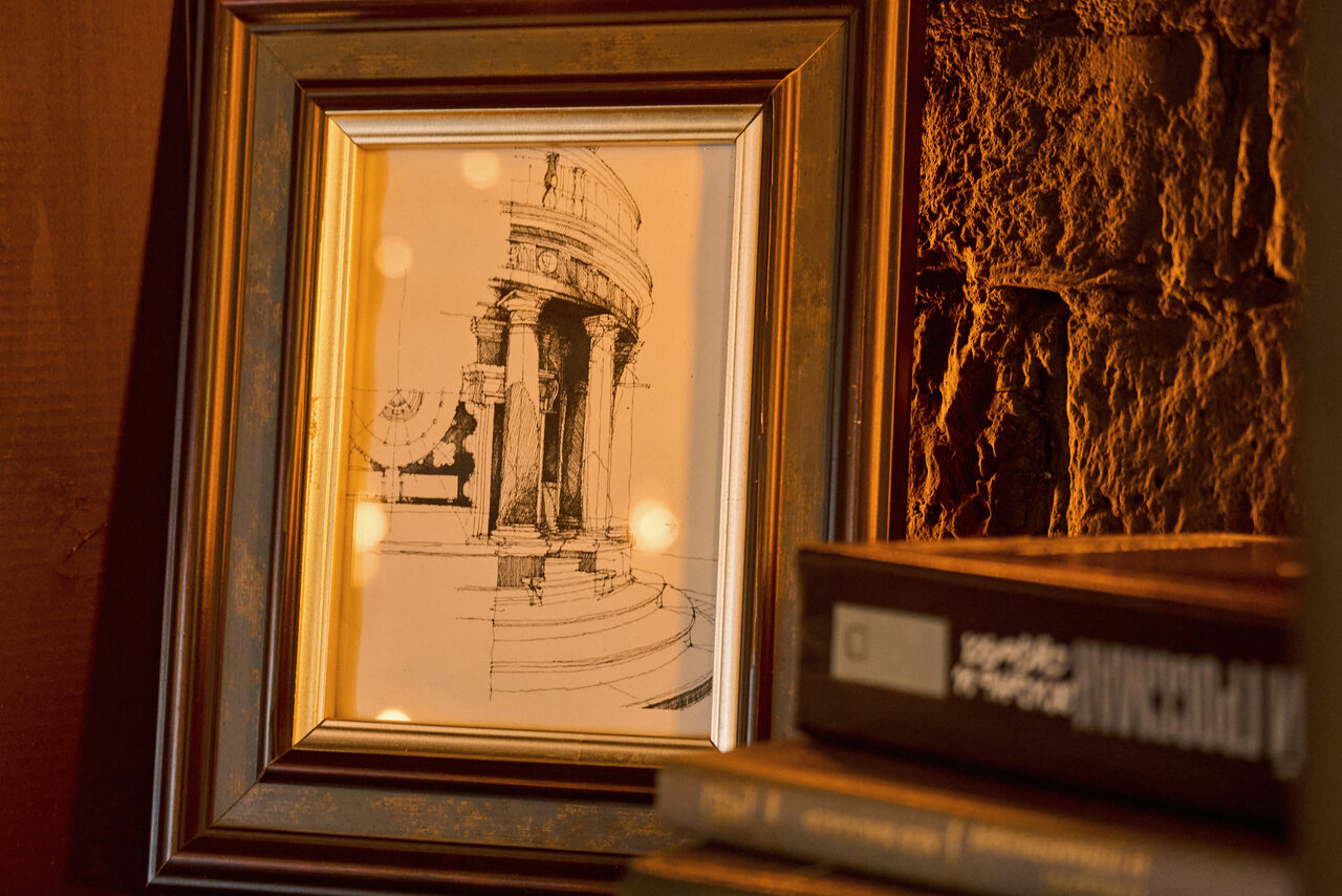 Арендовать Tangiers Lounge(Танжер Лаундж) . Картина в золотистой раме с изображением архитектурного сооружения на фоне текстурированной кирпичной стены. Рядом стопка книг с толстыми корешками. Теплое освещение.