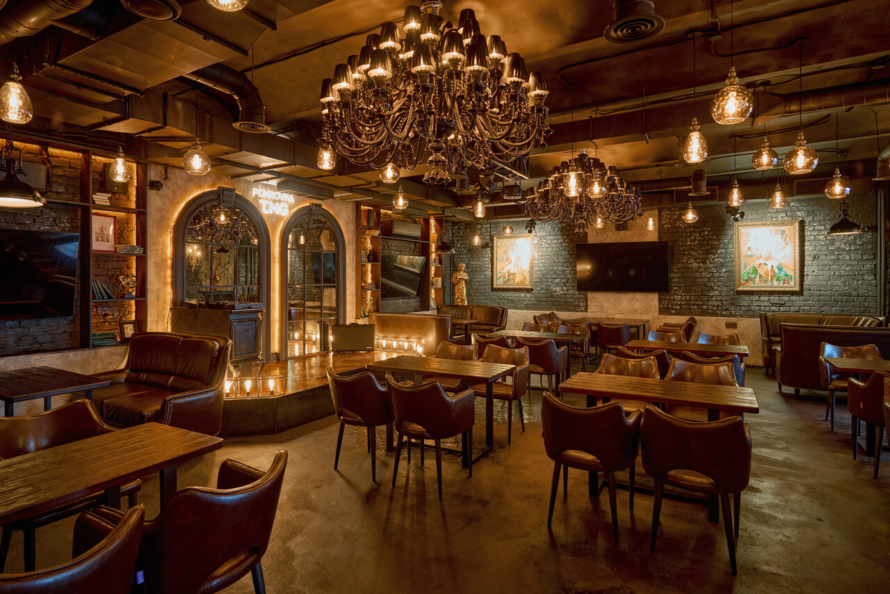 Арендовать Tangiers Lounge(Танжер Лаундж) . Ресторанное пространство с темными кожаными креслами и деревянными столами, просторная планировка. Стены из темного кирпича, черные люстры и лампочки создают теплое освещение. На стенах висят картины и телевизор. Архитектурные арки с зеркалами.