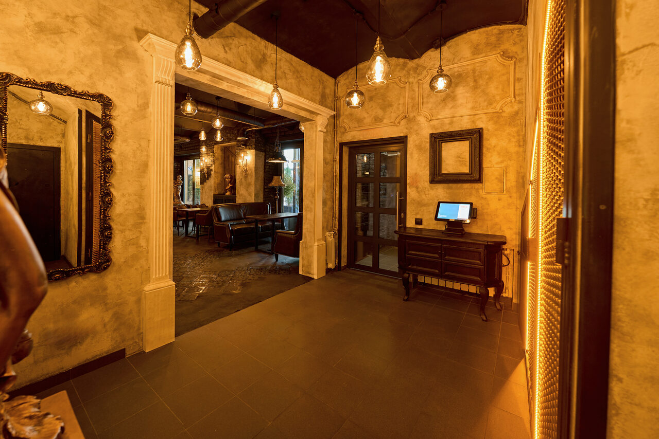 Арендовать Tangiers Lounge(Танжер Лаундж) . Коридор с арочным входом в зал. На стенах декоративная штукатурка. Подвесные лампы создают тёплое освещение. Справа стол с монитором. Слева зеркало. За аркой видны столы и кресла в тёмных тонах. Потолок тёмный.