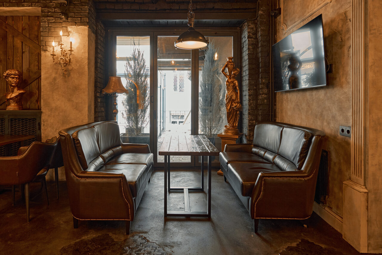 Арендовать Tangiers Lounge(Танжер Лаундж) . Пространство с двумя коричневыми кожаными диванами и деревянным столом между ними, на стене телевизор, рядом деревянная статуя, подвесной светильник и бра, окна с декоративными растениями, кирпичные стены и полированный бетонный пол.