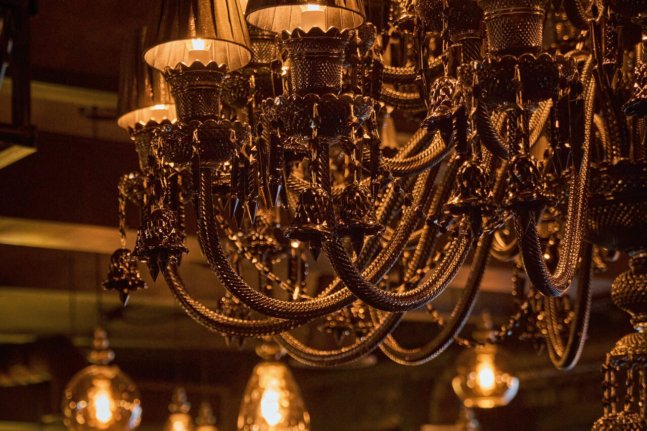 Арендовать Tangiers Lounge(Танжер Лаундж) . Большая декоративная люстра с множеством ламп и свисающих элементов. Тёмные металлические детали, тёплое золотистое освещение, создающее атмосферный полумрак. На заднем плане видны ещё несколько меньших светильников.