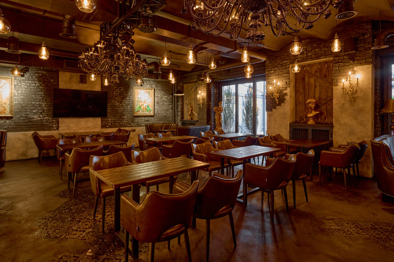 Арендовать Tangiers Lounge(Танжер Лаундж) . Просторное помещение с кирпичными стенами и винтажными люстрами. Кожаные стулья и деревянные столы расставлены рядами. Металлические светильники с теплым освещением. На стенах картины и бюсты. Темные тона интерьера, большие окна с занавесками.