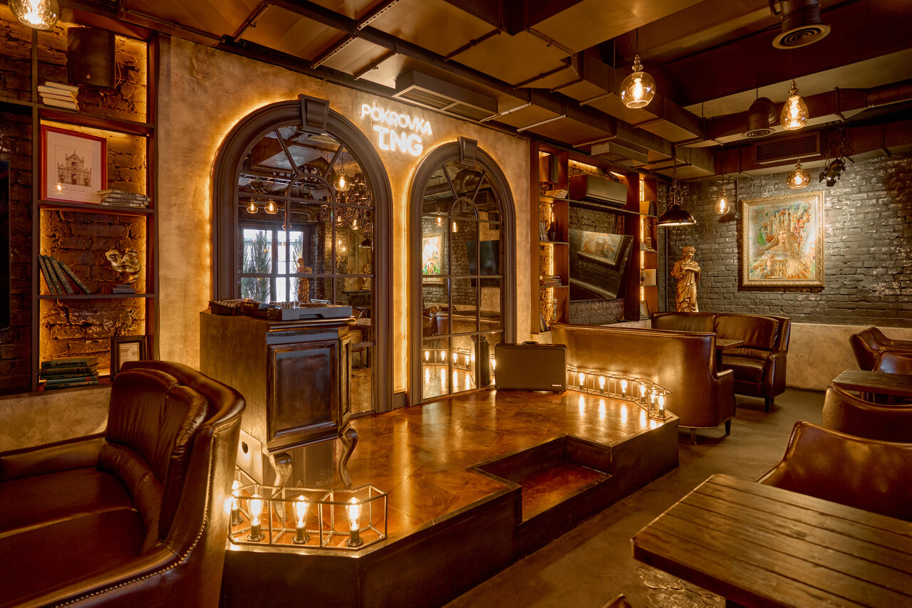 Арендовать Tangiers Lounge(Танжер Лаундж) . Лофт с кирпичными стенами и деревянными полками. Кожаные кресла и диваны, деревянные столы. Пол с подсветкой из свечей. Арочные двери, неоновая надпись. Потолок с открытыми балками и подвесными лампами. Настенная картина и статуэтка. Тёплое освещение.