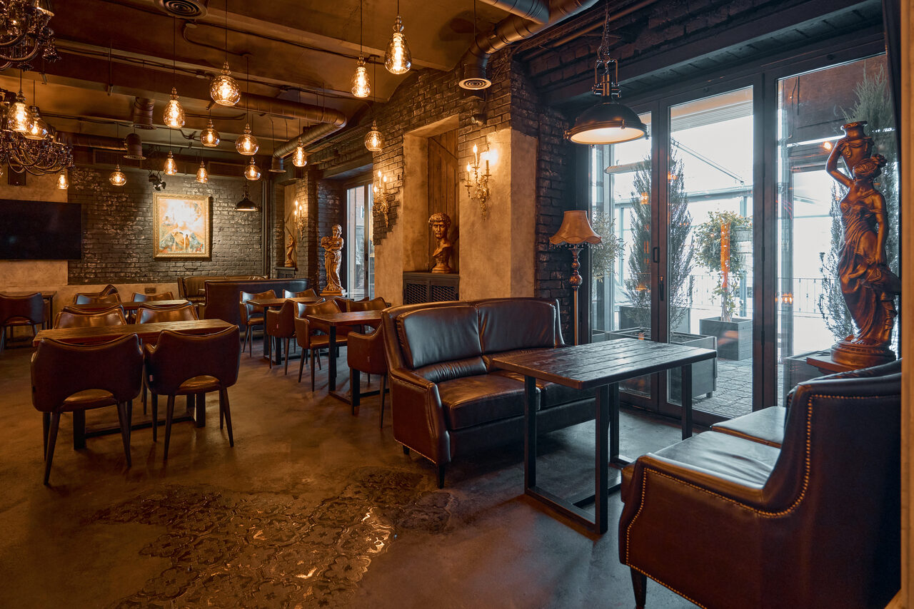 Арендовать Tangiers Lounge(Танжер Лаундж) . Лофт-пространство с бетонным полом и кирпичными стенами. Темная мебель: кожаные диваны, стулья, деревянные столы. Мягкое теплое освещение с лампами и бра. Декор в виде статуй и картин. Широкие окна с выходом на террасу, занавеси и растения в горшках.