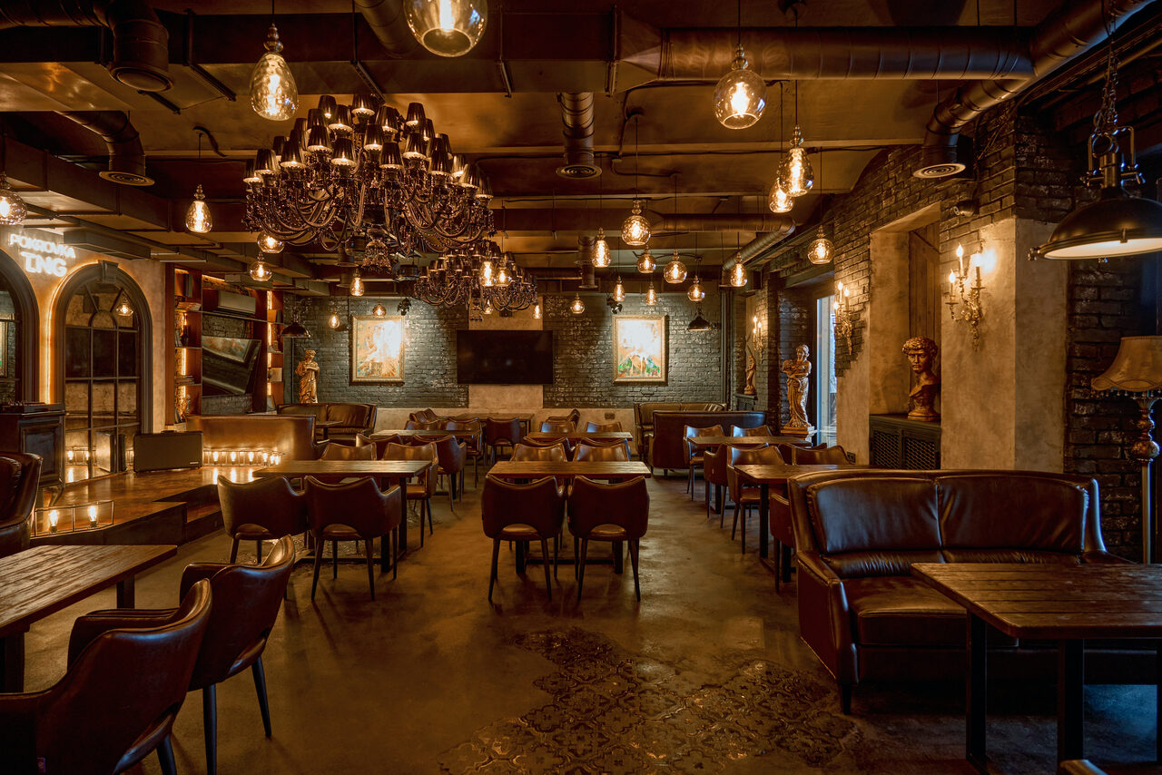 Арендовать Tangiers Lounge(Танжер Лаундж) . Просторный зал с кирпичными стенами и бетонным полом. Деревянные столы и кожаные стулья. Потолочные светильники и большой люстр с лампочками. Окна частично закрыты шторами. На стенах картины и декоративные элементы. Пастельные и тёмные оттенки.