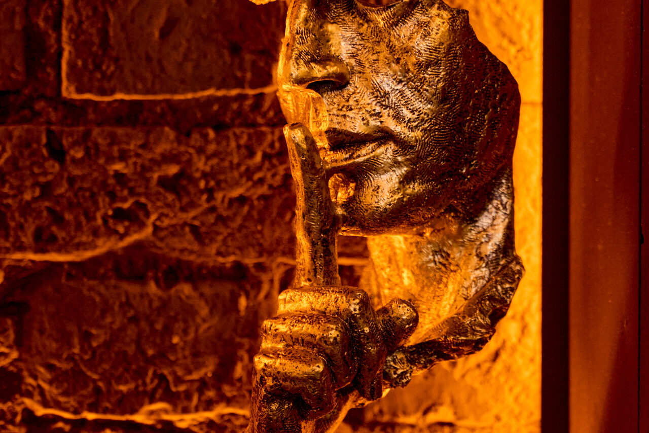 Арендовать Tangiers Lounge(Танжер Лаундж) . Скульптура из металла в виде лица с прикладывающим палец к губам на фоне кирпичной стены с тёплым освещением в оттенках оранжевого.