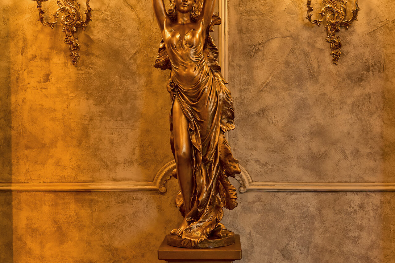 Арендовать Tangiers Lounge(Танжер Лаундж) . Золотая статуя в классическом стиле с изображением женщины стоит на постаменте перед текстурированной стеной теплого коричневого оттенка. Справа и слева позолоченные декоративные элементы с орнаментом. Освещение теплое.