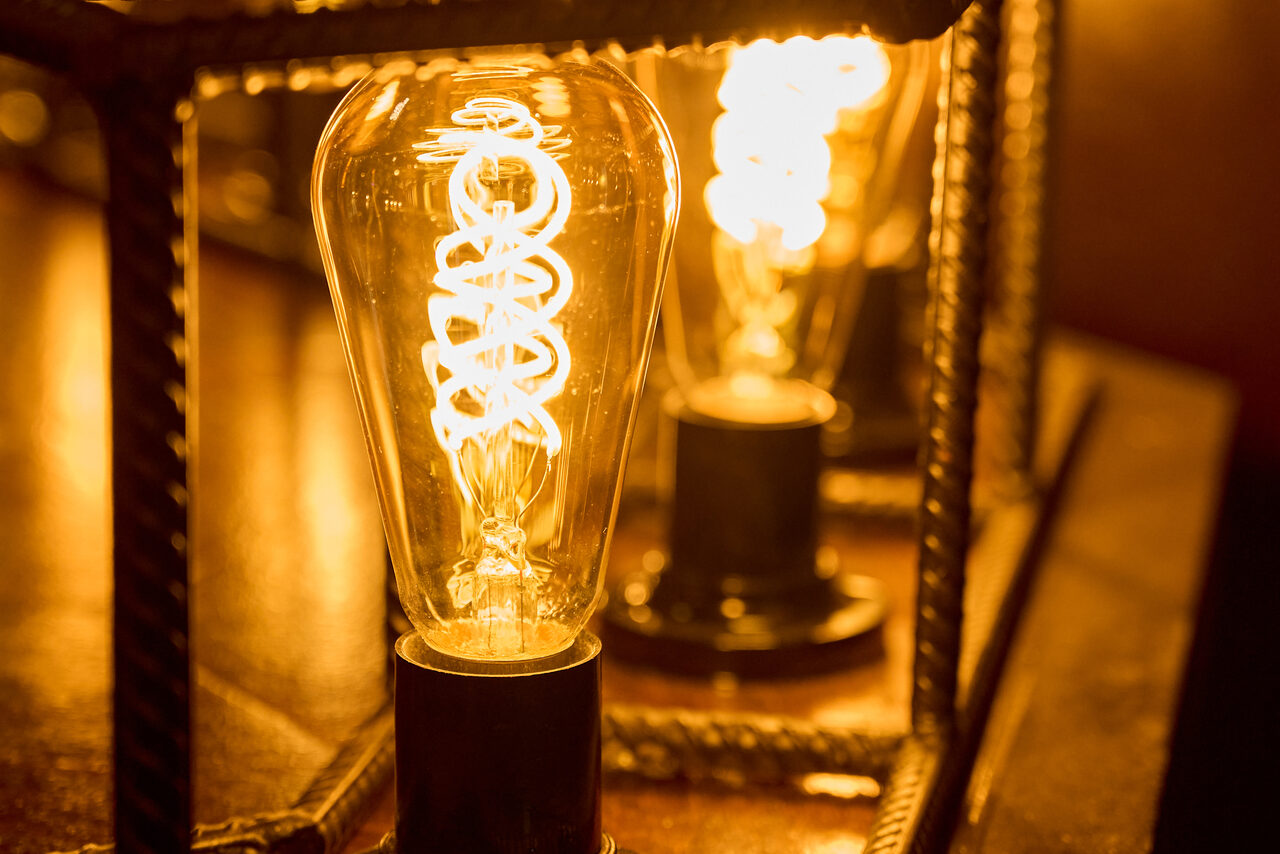 Арендовать Tangiers Lounge(Танжер Лаундж) . Включенная лампа с нитью накаливания в металлическом каркасе на деревянной поверхности, отражающаяся в зеркале. Тёплое жёлтое освещение, металлические элементы с рельефной текстурой.