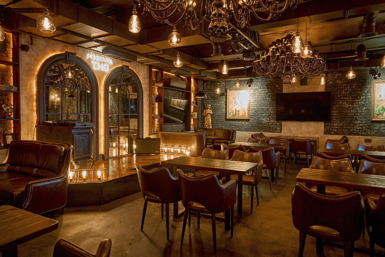 Арендовать Tangiers Lounge(Танжер Лаундж) . Ресторанное пространство с деревянными столами и кожаными стульями, тёмные кирпичные стены, подвесные светильники и люстры, стильные арочные входы, барная стойка. Экраны и картины на стенах. Теплое освещение.