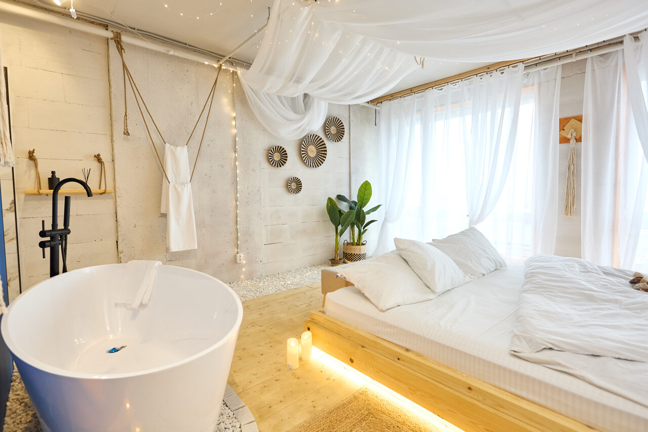 Арендовать Пляжный лофт с галькой, открытой ванной, панорамными окнами. Спальня со светлыми стенами и потолком с белым декором, большая кровать с белым постельным бельём, отдельно стоящая белая ванна, чёрный смеситель, декоративные тарелки на стене, зелёное растение в горшке, свечи, деревянный пол.