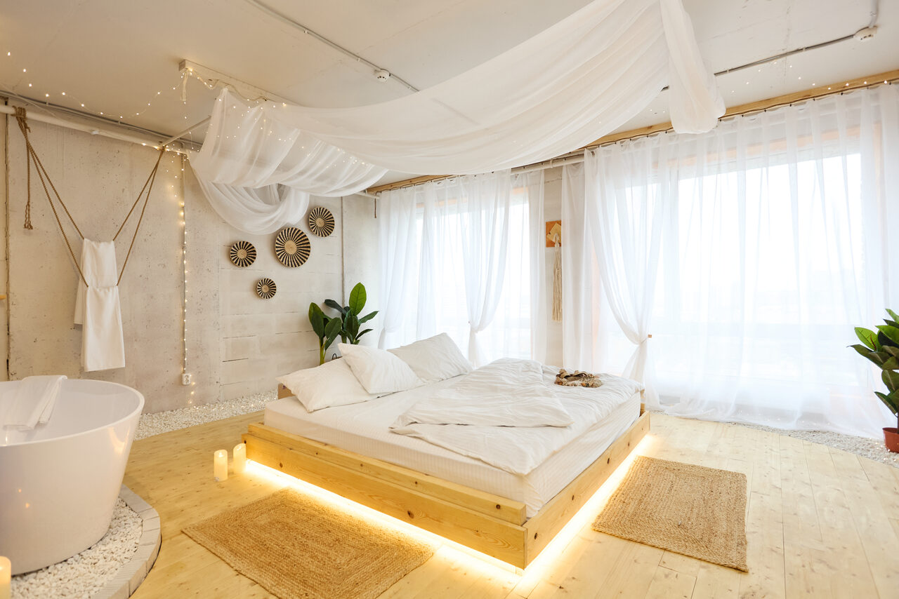 Арендовать Пляжный лофт с галькой, открытой ванной, панорамными окнами. Пространство с деревянным полом и светлыми стенами. В центре кровать на подиуме с белыми покрывалами и подушками. Справа ванна округлой формы, рядом дорожки из ковров. Большие окна с белыми занавесками. Украшение на стене и несколько горшков с растениями.