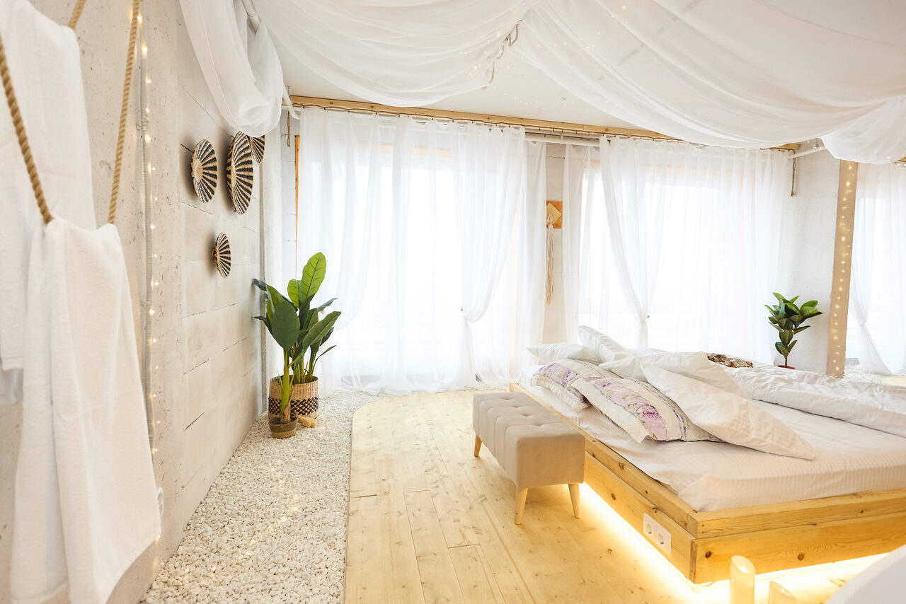 Арендовать Пляжный лофт с галькой, открытой ванной, панорамными окнами. Светлая спальня с кроватью на деревянном подиуме, фоновое освещение. Белые занавески на окнах, растения в горшках, серая скамья у изножья кровати. Стены украшают декоративные элементы, пол из светлого дерева и белых камней.