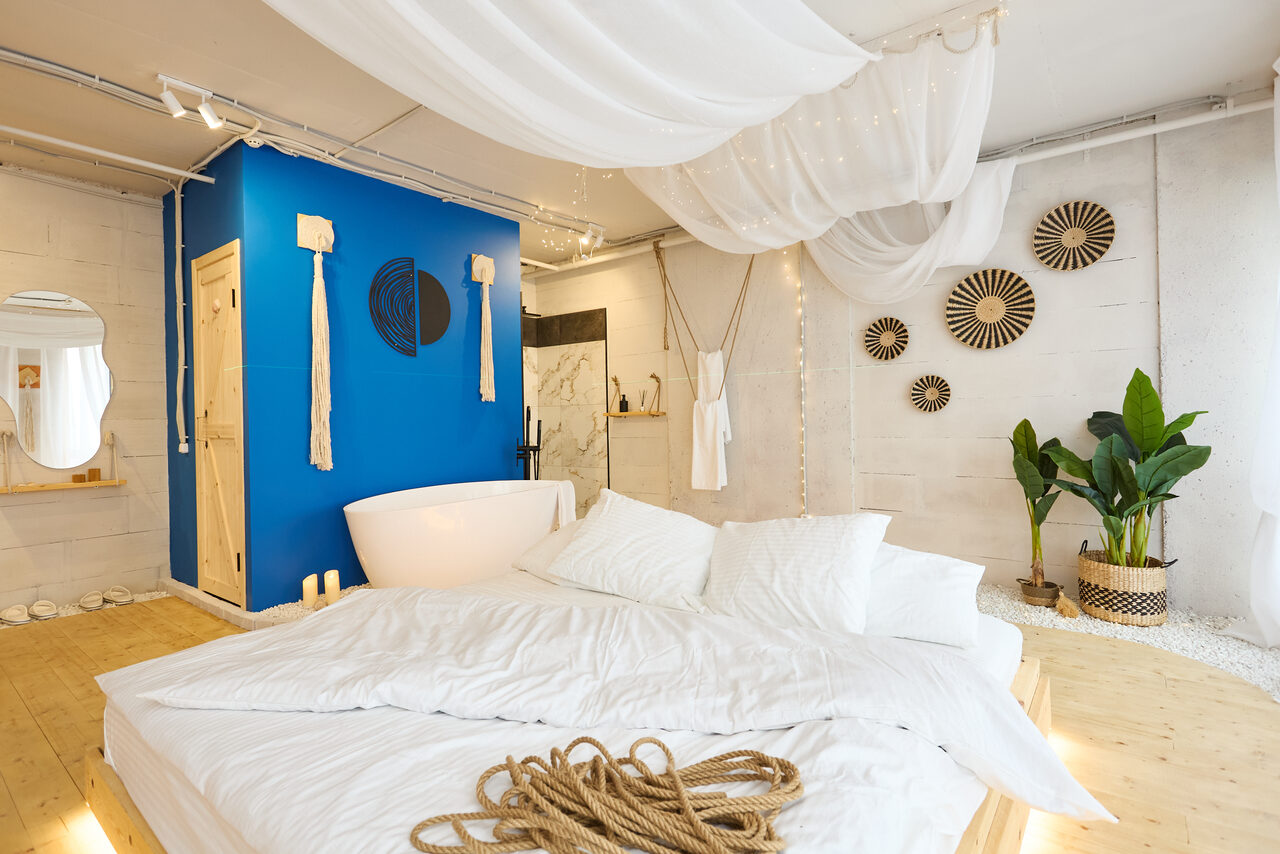 Арендовать Пляжный лофт с галькой, открытой ванной, панорамными окнами. Спальня с большой кроватью и белым постельным бельём. Белые стены и деревянный пол, стены украшены декором. Ванна в комнате возле ярко-синей стены. Зелёные растения в углу. Белые тканевые драпировки на потолке.