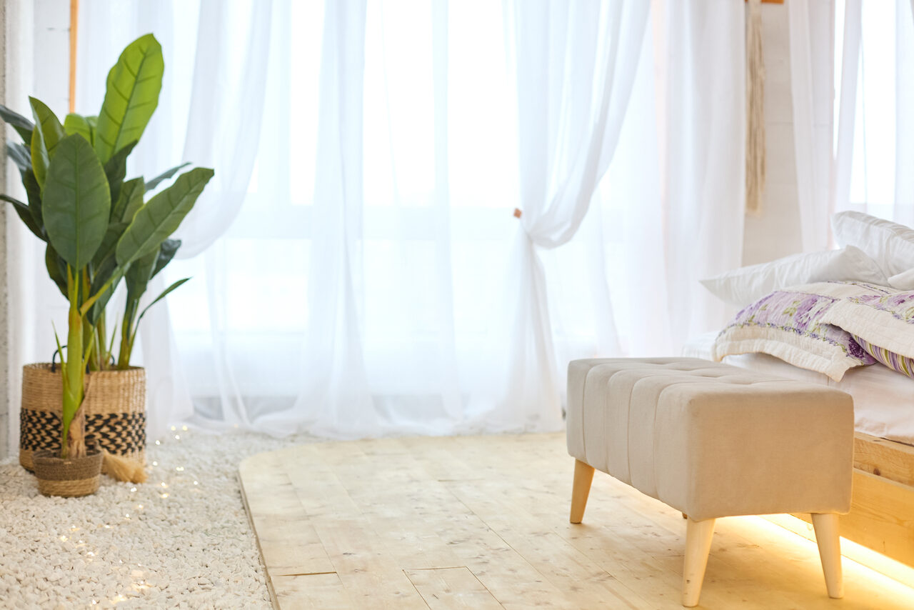 Арендовать Пляжный лофт с галькой, открытой ванной, панорамными окнами. Светлая комната с деревянным полом и белыми прозрачными шторами. Мягкая скамья с бежевой обивкой у изножья кровати. По бокам зелёные растения в плетёных корзинах. Светлое постельное бельё, подушки с узором сиреневых оттенков.