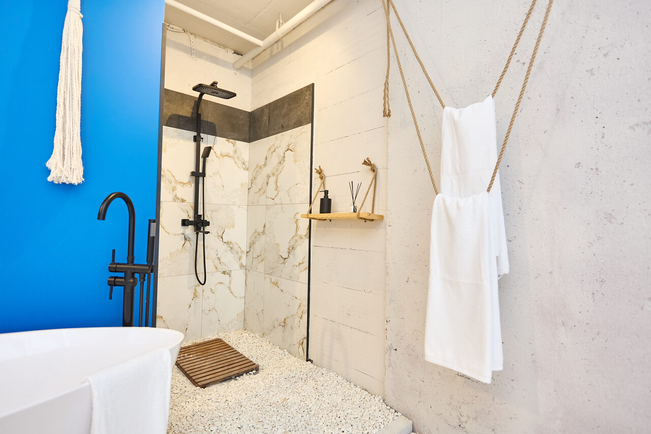 Арендовать Пляжный лофт с галькой, открытой ванной, панорамными окнами. Ванная комната с открытым душем, черный душевой гарнитур, бежевые мраморные плитки, белый гравий на полу. Белоснежные полотенца висят на веревках, ванна с черным смесителем, синий акцент на стене, деревянная полочка с аксессуарами.