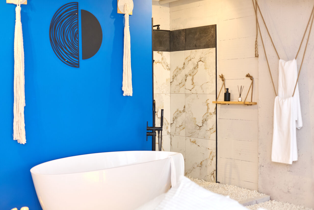 Арендовать Пляжный лофт с галькой, открытой ванной, панорамными окнами. Ванная комната с белой овальной ванной на фоне ярко-синей стены с декоративными элементами. Светло-серые плитки под мрамор, деревянная полка с декором. Мягкое освещение, белые полотенца на веревке.