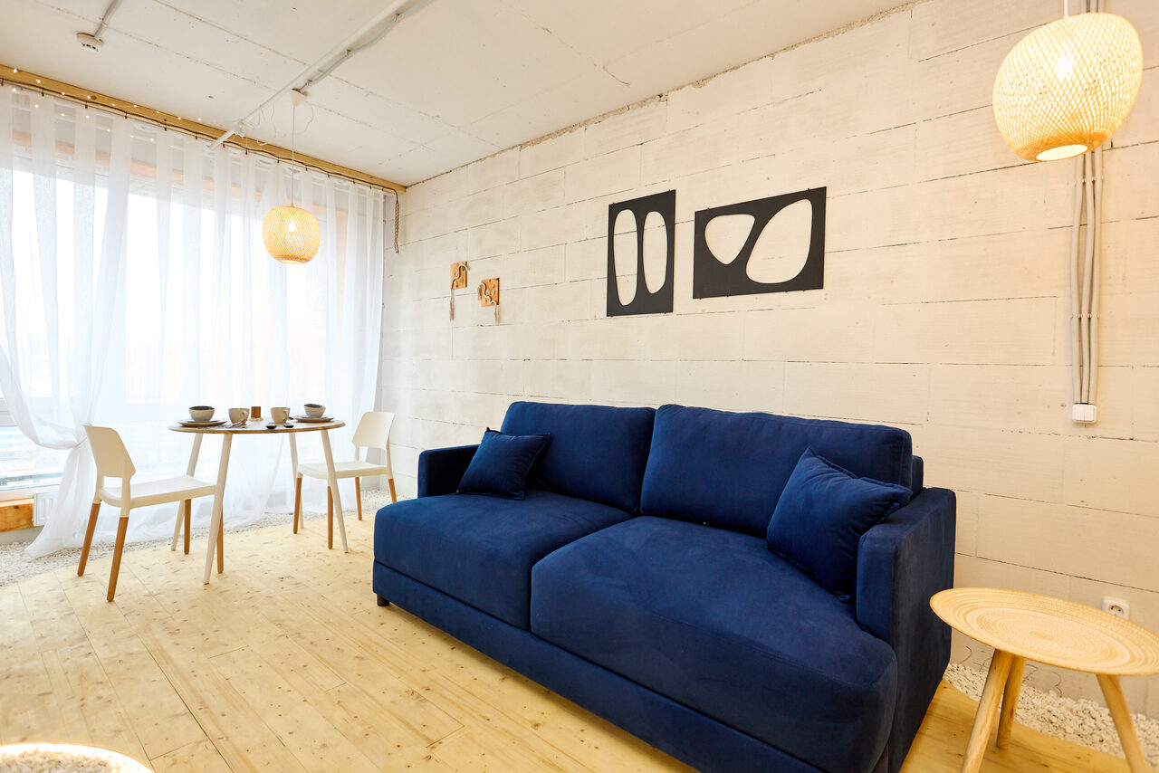 Арендовать Пляжный лофт с галькой, открытой ванной, панорамными окнами. Меблированная гостиная синим диваном и круглыми подушками на светлом деревянном полу. На стене декоративные элементы в черной рамке. Белый обеденный стол с четырьмя креслами. Большое окно с белой шторой. Подвесные светильники из светлого материала.