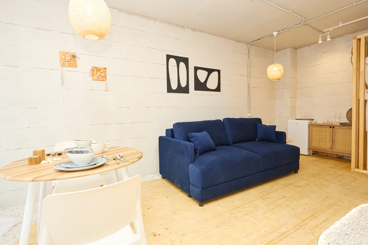 Арендовать Пляжный лофт с галькой, открытой ванной, панорамными окнами. Комната с белыми бетонными стенами, синим диваном, круглым деревянным столом и двумя белыми стульями. На столе посуда. Подвесные светильники, деревянный пол, небольшой холодильник. Настенные абстрактные картины.