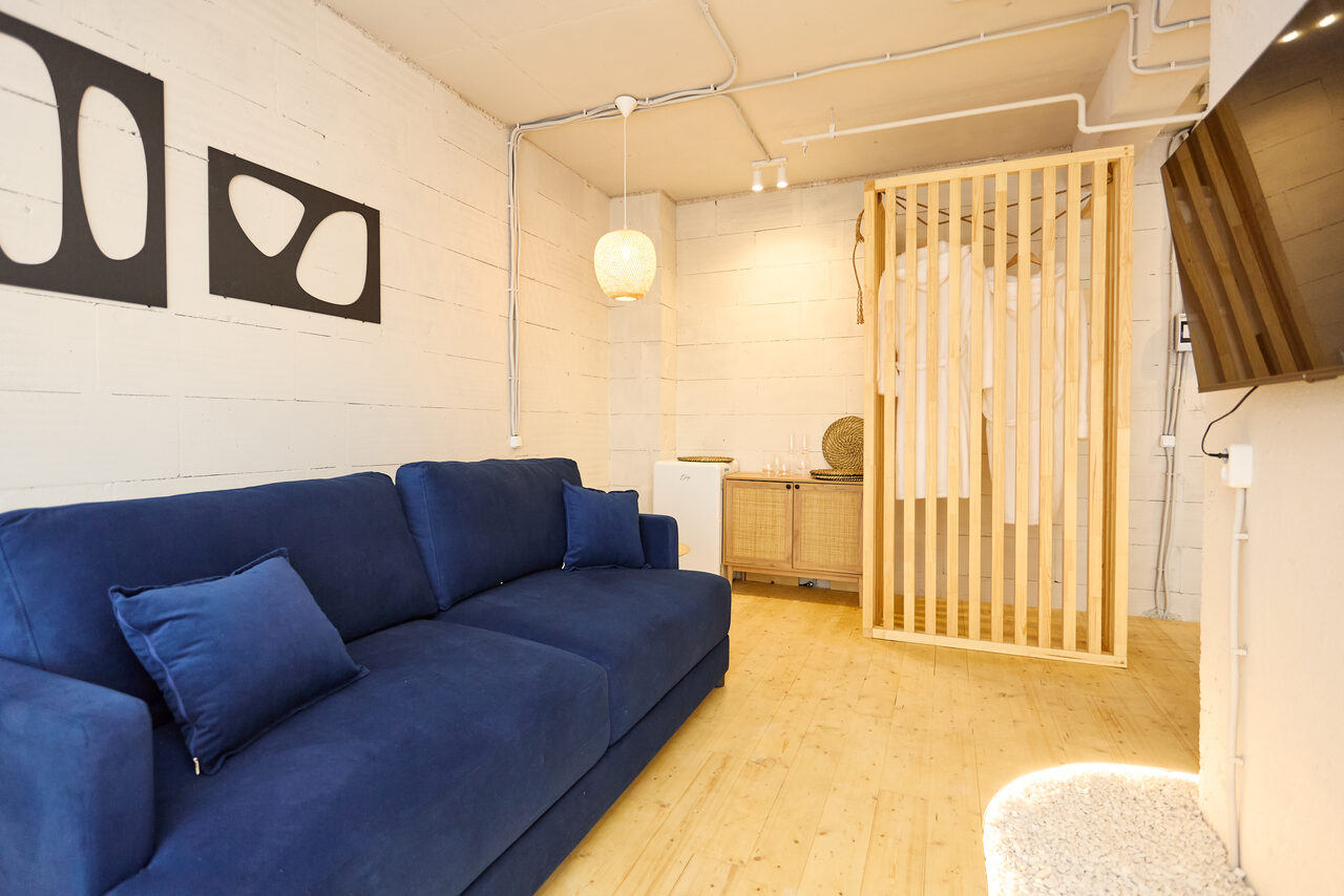 Арендовать Пляжный лофт с галькой, открытой ванной, панорамными окнами. Просторная комната с деревянным полом, белыми стенами и открытым потолком. Синяя диванная зона, светлые деревянные перегородки, навесной светильник, настенный телевизор. Комод с декоративными элементами. Современное освещение с точечными светильниками.