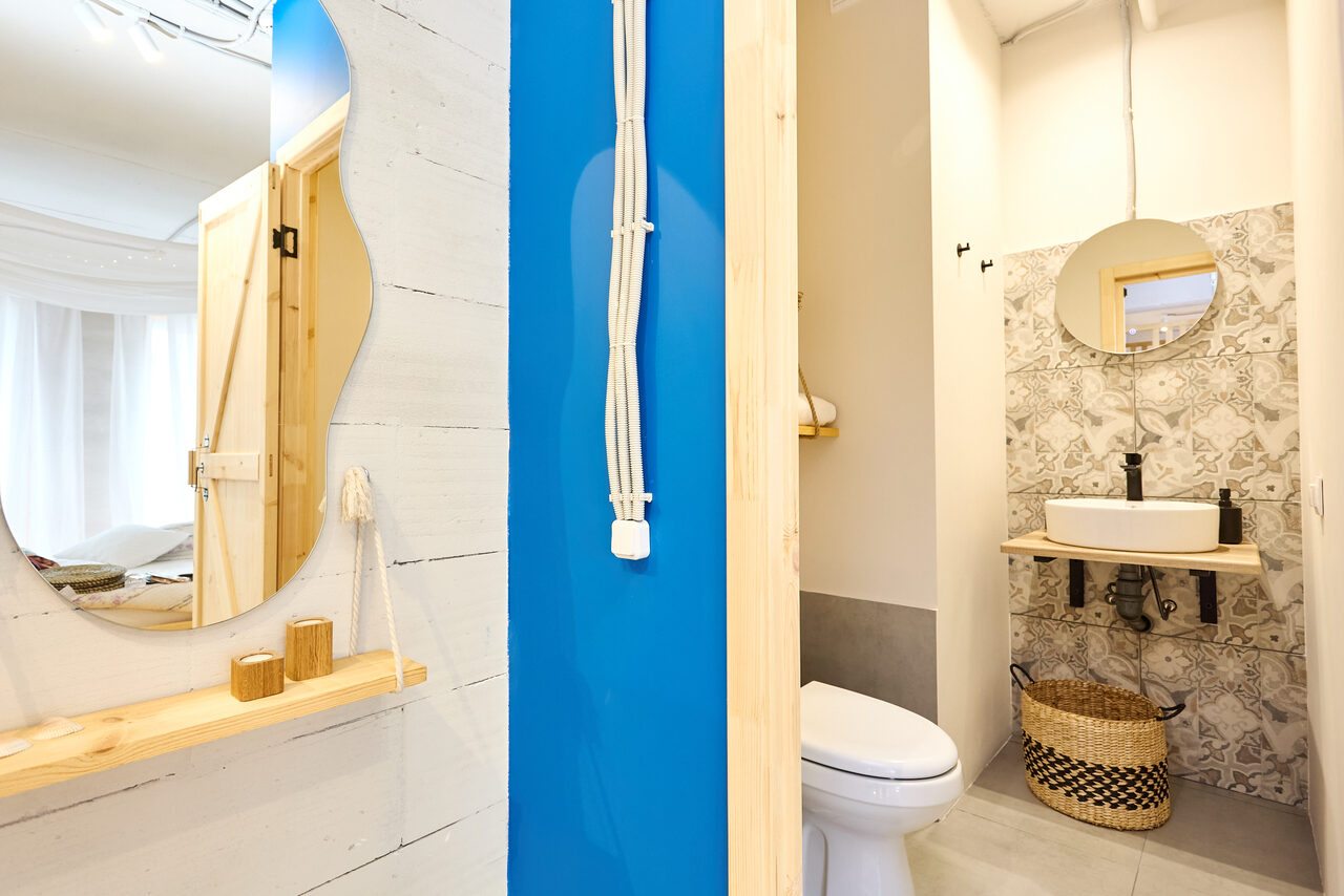 Арендовать Пляжный лофт с галькой, открытой ванной, панорамными окнами. Туалет с круглым зеркалом, раковиной, плетёной корзиной и унитазом; стены из белой и синей плитки, белые стены, светлое деревянное обрамление; деревянная полка с кружевным зеркалом; яркое освещение.