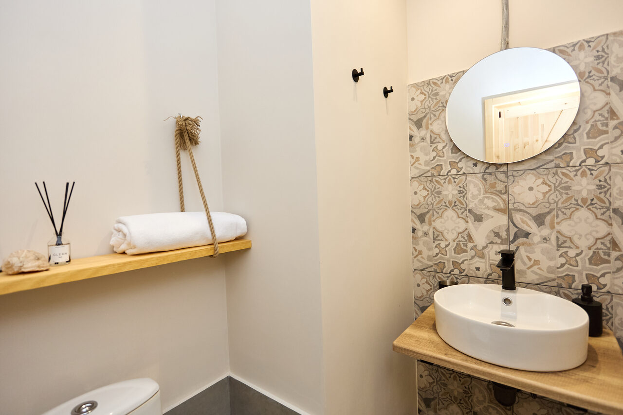 Арендовать Пляжный лофт с галькой, открытой ванной, панорамными окнами. Ванная комната с круглым зеркалом над белой раковиной на деревянной столешнице. Стена с плиткой в узорах, открытая полка с полотенцем и декором. Белые стены, металлические крючки, нейтральное освещение.