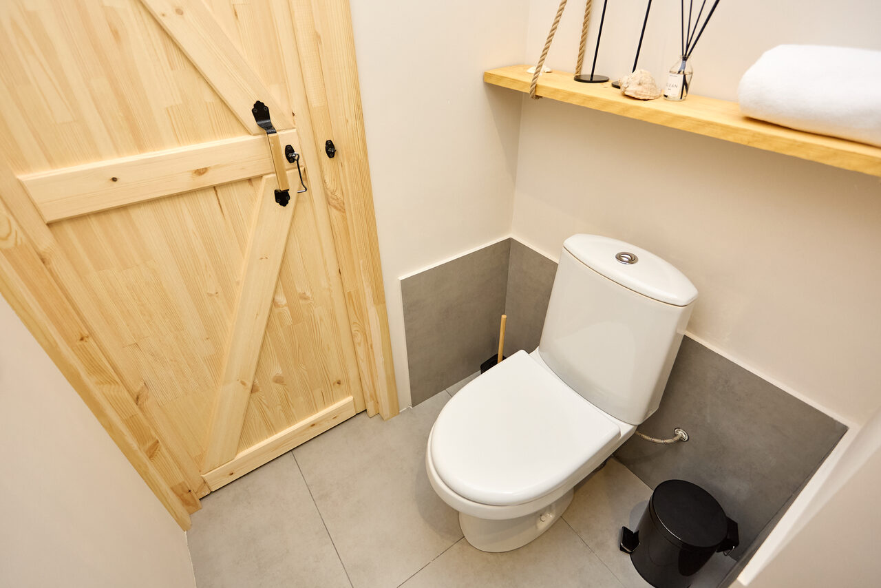 Арендовать Пляжный лофт с галькой, открытой ванной, панорамными окнами. Туалетное пространство с унитазом, дверью из светлого дерева, полками с декоративными элементами. Пол и часть стен облицованы серыми плитками. Светлый верх стен. Черный мусорный бак и щетка для туалета.