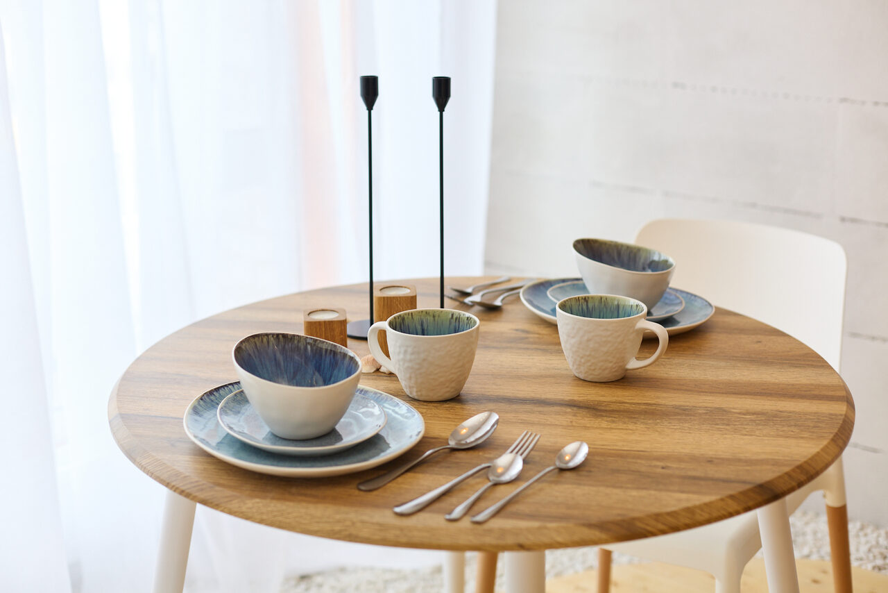 Арендовать Пляжный лофт с галькой, открытой ванной, панорамными окнами. Круглый деревянный стол с тарелками и чашками, сервированными для трёх персон. На столе столовые приборы, две высокие чёрные свечи. Стулья белого цвета, пол светлый. Фоновая стена светлая с текстурой. Естественное освещение через белую занавеску.