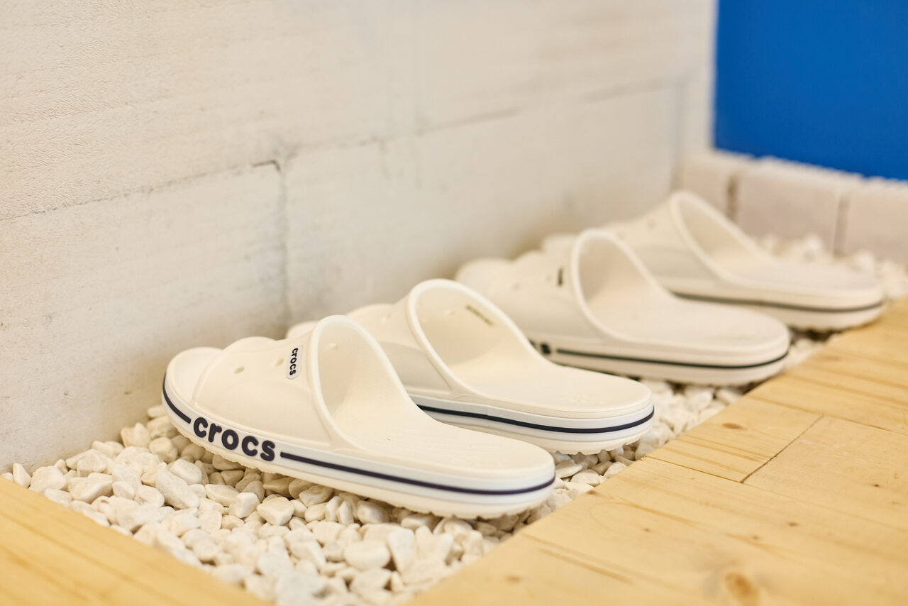 Арендовать Пляжный лофт с галькой, открытой ванной, панорамными окнами. Пол белого цвета из небольших камней вдоль светлой кирпичной стены, на которых находятся три пары белых шлёпанцев с надписью. Справа часть деревянного настила и синий цвет на боковой стене.