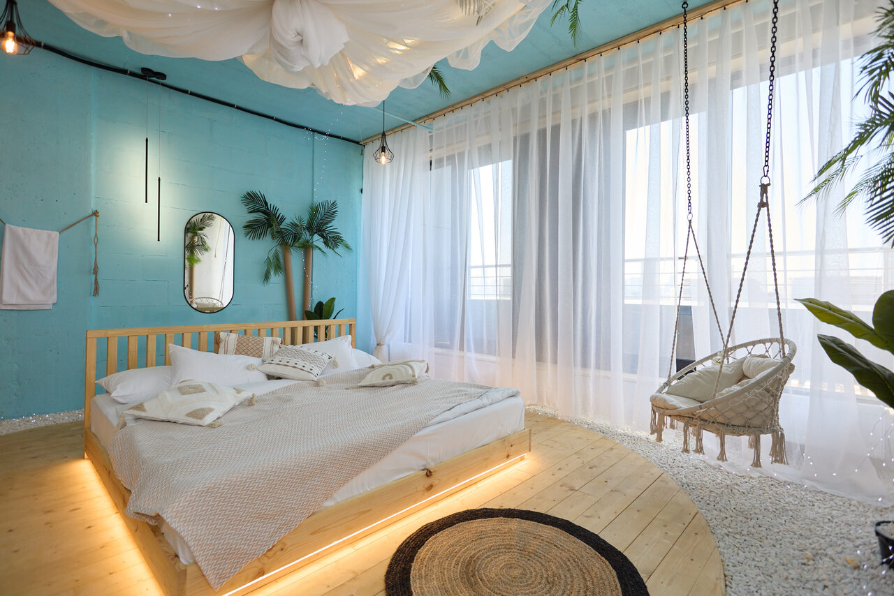 Арендовать Лофт Мальдивы с открытым душем, балконом и видом на Сити. Спальня с голубыми стенами, кровать с деревянным каркасом, подвесное кресло, пол из светлого дерева, прозрачные шторы, декоративные растения, подвесные лампы, овальное зеркало, белый текстиль на кровати, плетеный ковер.