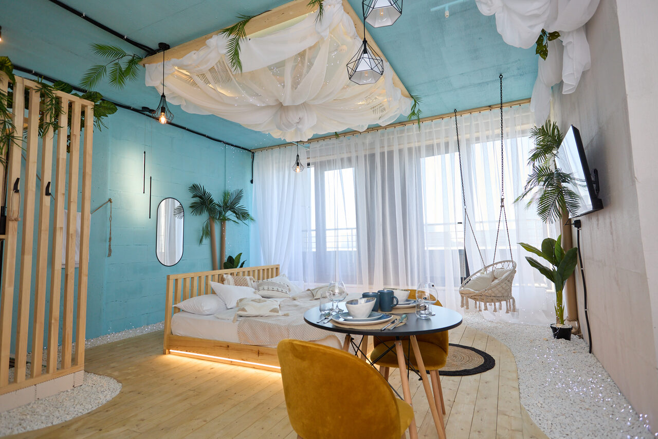 Арендовать Лофт Мальдивы с открытым душем, балконом и видом на Сити. Спальня с бирюзовыми стенами, кроватью под балдахином, деревянным полом. Обеденный стол с желтыми стульями, балкон с прозрачными шторами. Подвесное кресло, множество растений, телевизор на стене. Декоративные элементы из ткани и геометрические светильники.