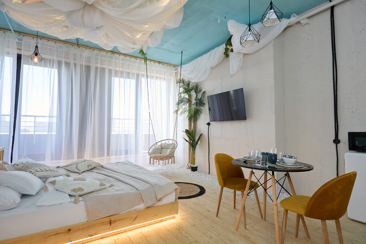Арендовать Лофт Мальдивы с открытым душем, балконом и видом на Сити. Спальня с деревянным полом, низкой кроватью, двумя желтыми стульями и круглым обеденным столом. Настенный телевизор, подвесное освещение, белые занавески на окнах, голубой потолок с драпировкой. Декоративные растения и подвесное кресло.