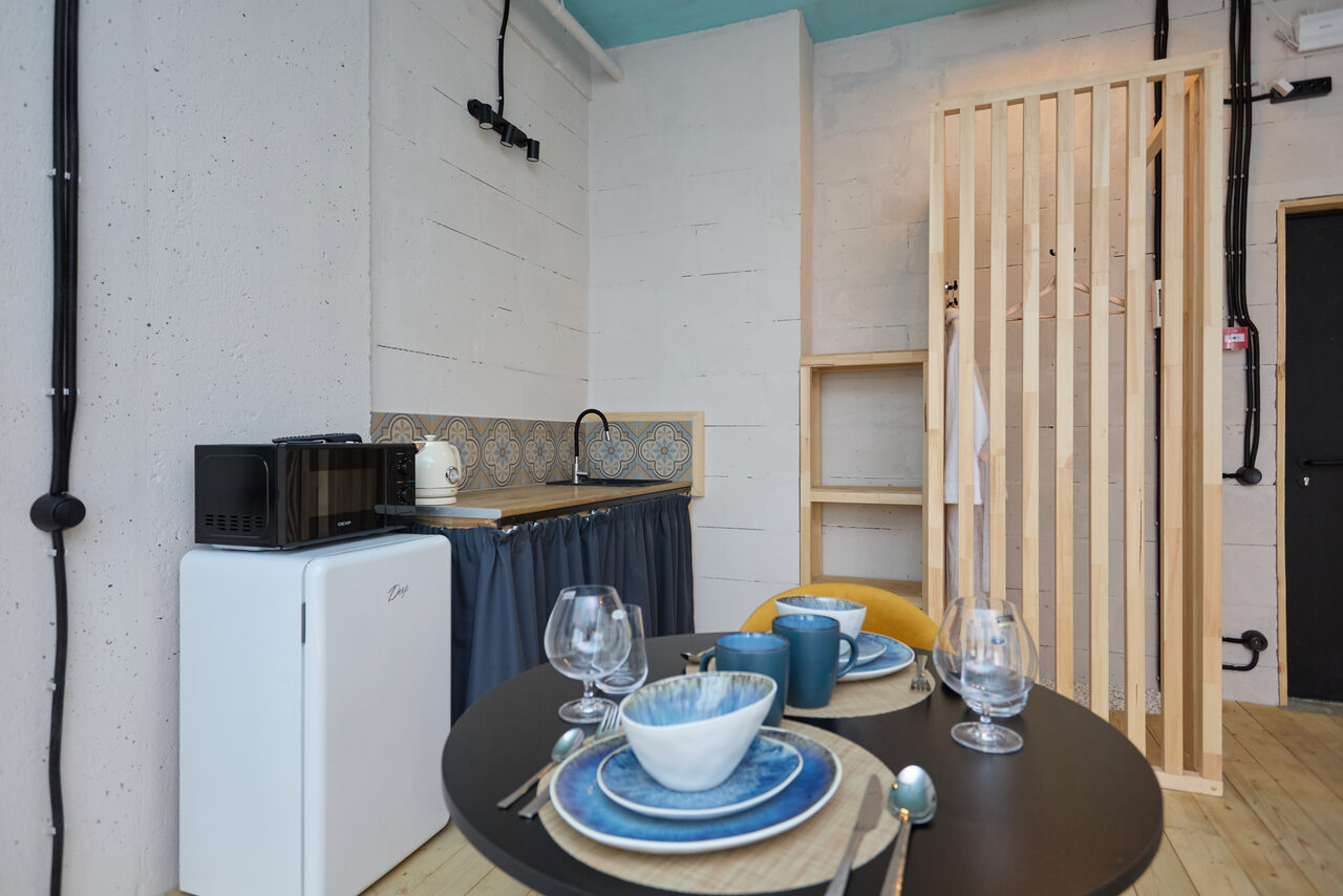 Арендовать Лофт Мальдивы с открытым душем, балконом и видом на Сити. Маленькая кухонная зона со столом и двумя стульями. На столе три тарелки, чашки и бокалы. В углу мини-холодильник и микроволновка. Раковина с узорчатым фартуком. Слева от раковины текстильная штора. Светлые стены и деревянная перегородка.