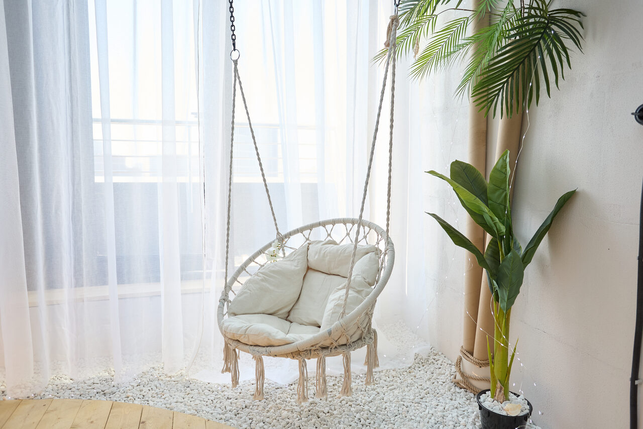 Арендовать Лофт Мальдивы с открытым душем, балконом и видом на Сити. Круглое подвесное кресло с подушками на веревочных цепях, белые шторы от потолка до пола, декоративные белые камни на полу, деревянное половое покрытие, зеленое растение в горшке, белая стена.
