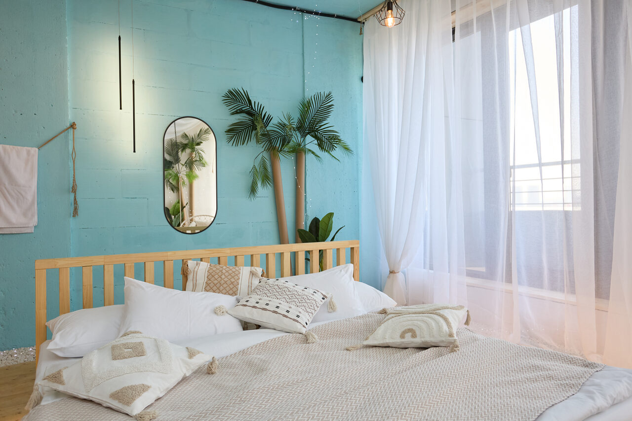 Арендовать Лофт Мальдивы с открытым душем, балконом и видом на Сити. Спальня с кроватью и подушками, деревянная спинка, светло-голубая стена с зеркалом овальной формы, искусственные пальмы, белые прозрачные шторы, окно, регулируемое освещение на стене, деревянный пол.