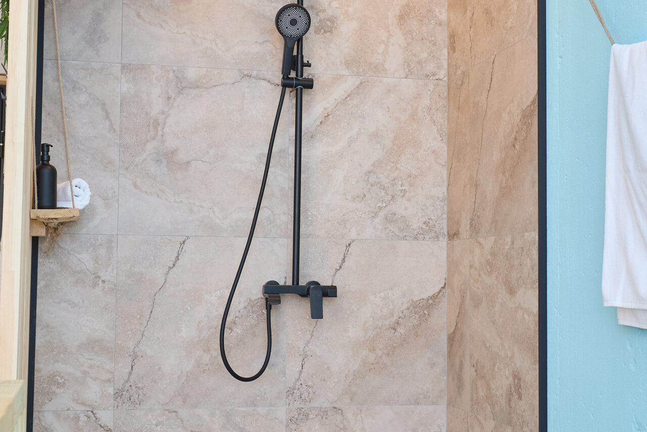 Арендовать Лофт Мальдивы с открытым душем, балконом и видом на Сити. Душевая зона с черным набором душа на стене, облицованной крупной плиткой бежевого цвета с мраморным узором. Слева небольшая полка с черным флаконом и белым полотенцем. Справа белое полотенце на голубой стене.