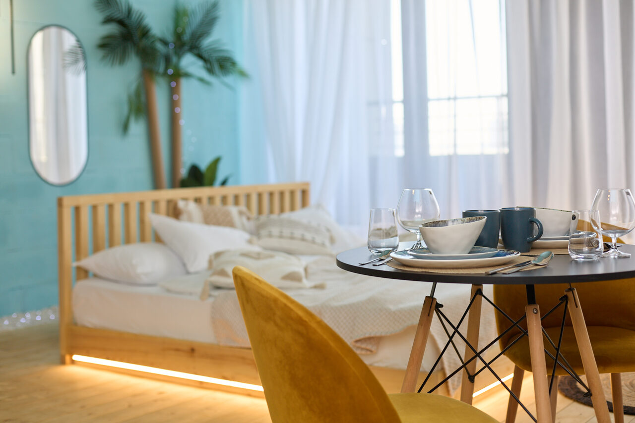 Арендовать Лофт Мальдивы с открытым душем, балконом и видом на Сити. Спальня с голубыми стенами, кровать с постельным бельем, круглый обеденный стол с сервировкой, желтые стулья, зеркало, комнатное растение, белые шторы на окне, светлое деревянное покрытие на полу.