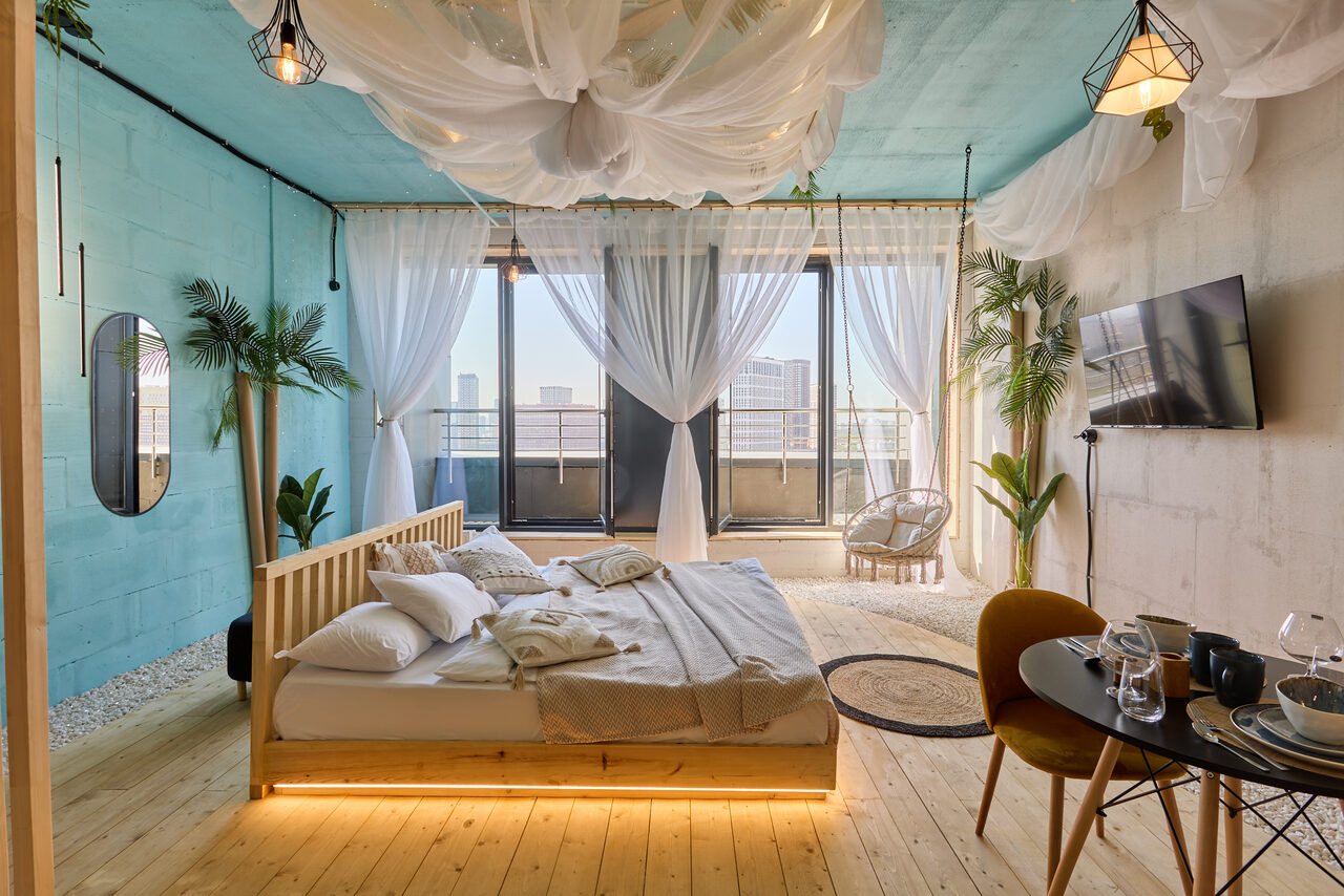 Арендовать Лофт Мальдивы с открытым душем, балконом и видом на Сити. Спальня с деревянным полом и потолком голубого цвета. Двуспальная кровать с подушками и покрывалом, рядом обеденный стол с посудой. Большие окна со светлыми шторами и видом на балкон. Растения в горшках, телевизор на стене, подвесные светильники.