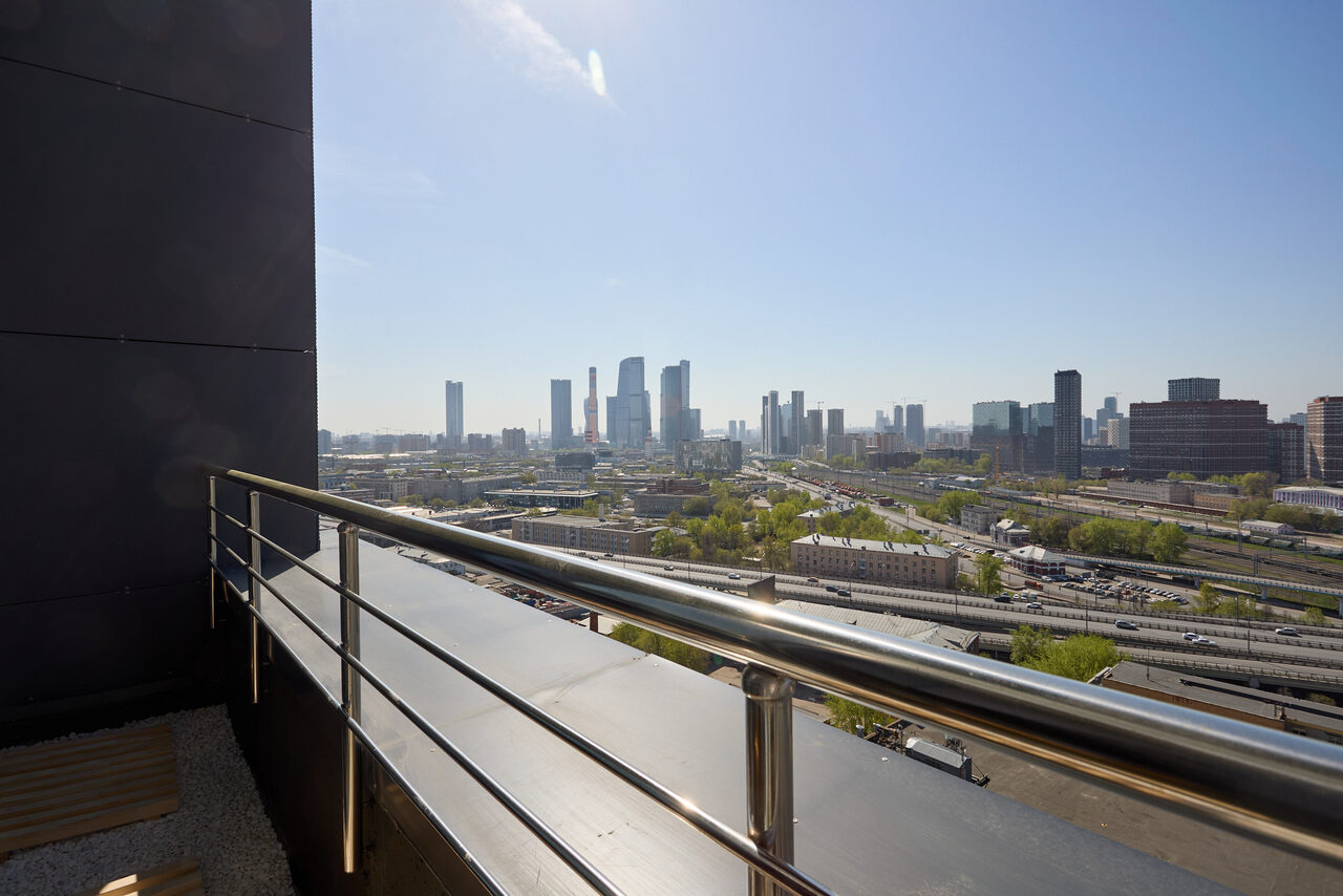 Арендовать Лофт Мальдивы с открытым душем, балконом и видом на Сити. Открытая терраса с перилами из металла, вид на городской ландшафт с небоскрёбами на горизонте. Чистое голубое небо и дневное освещение. Стены тёмного цвета, на полу гравий.