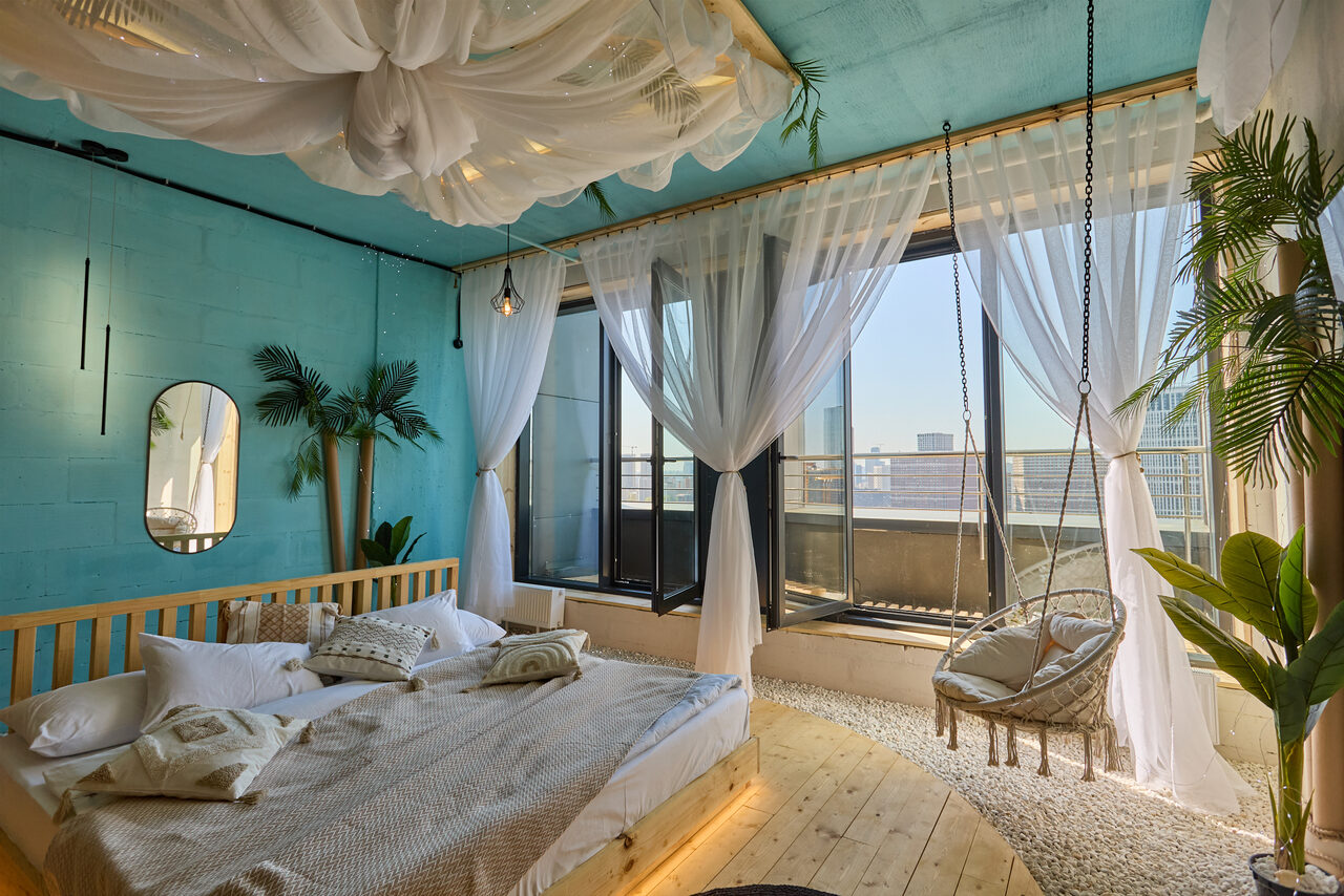 Арендовать Лофт Мальдивы с открытым душем, балконом и видом на Сити. Спальня с голубыми стенами и деревянным полом. Большая кровать с подушками, подвесное кресло, панорамные окна с белыми шторами. Подвесной декор на потолке, зеркала, зеленые растения, светлая мебель, светильники на стене.